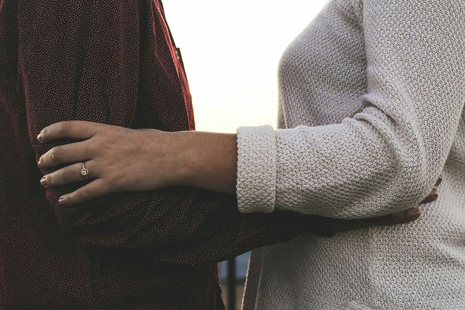 A couple, holding each other. One wearing a brown long sleeve sweater. One wearing a grey long sleeve sweater and wedding ring.